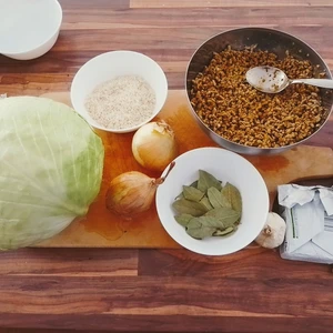 Küchenarbeitsplatte, auf dem ein Holzbrett liegt und auf welchem die Zutaten liegen. Ein Kohlkopf, Schüssel mit Lorbeerenblättern, Reis, eine Schüssel mit Hack, Margarine, Tomatenmark, Zwiebel, Knoblauch