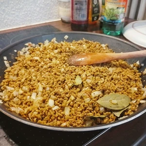 Pfanne mit angebratenen Hack und Zwiebel. Einzelne Lorbeerenblätter liegen in der Pfanne und ein Holzlöffel liegt auf dem Inhalt